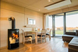 A modern living room with a wood-burning stove, a dining area with chairs, and a large window offering a view. - Holiday home with 2 bedrooms (Schwedeneck)
