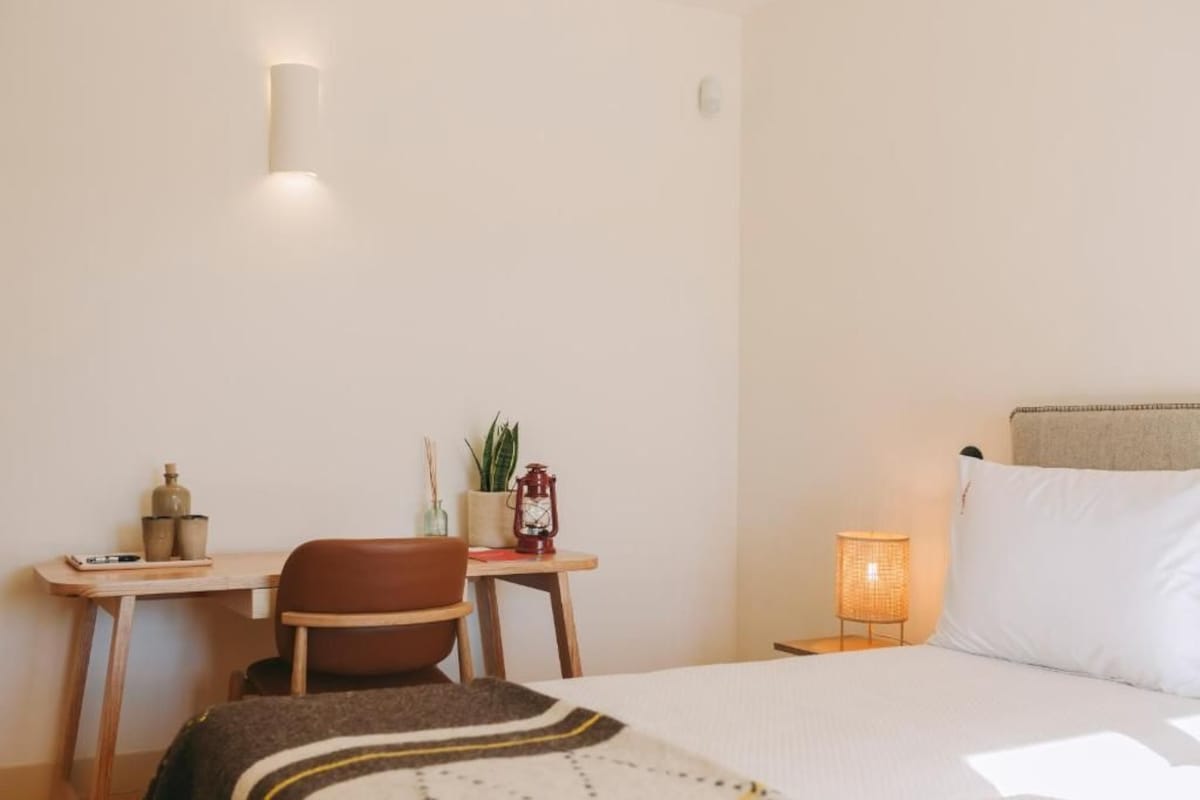 A bedroom with a bed, a desk, a chair, a nightstand with a lamp, and a wall-mounted light fixture.