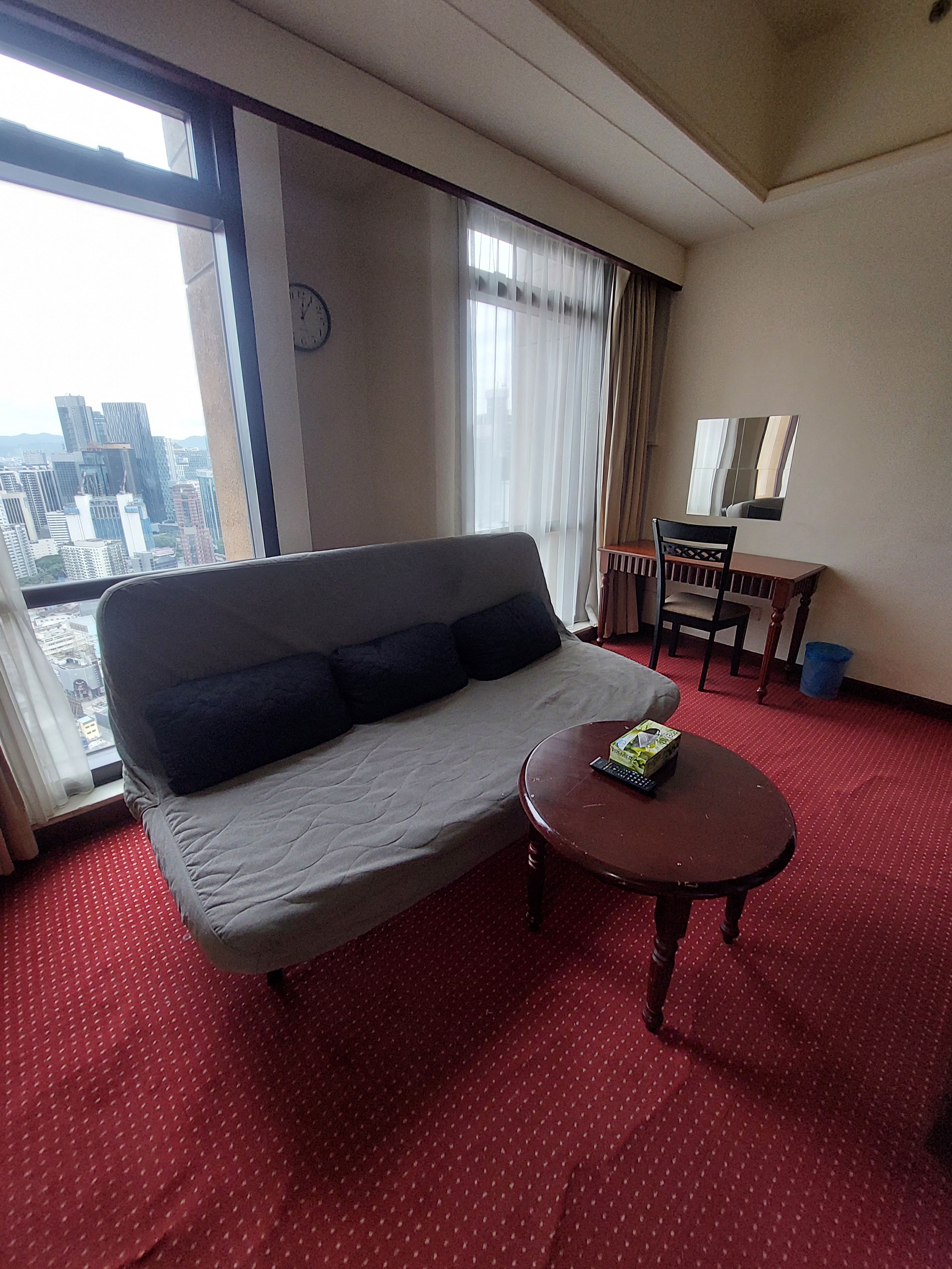 Ein Hotelzimmer mit einer Couch, einem kleinen Tisch, einem Sessel, einem Fenster mit Stadtblick und einer Wanduhr.