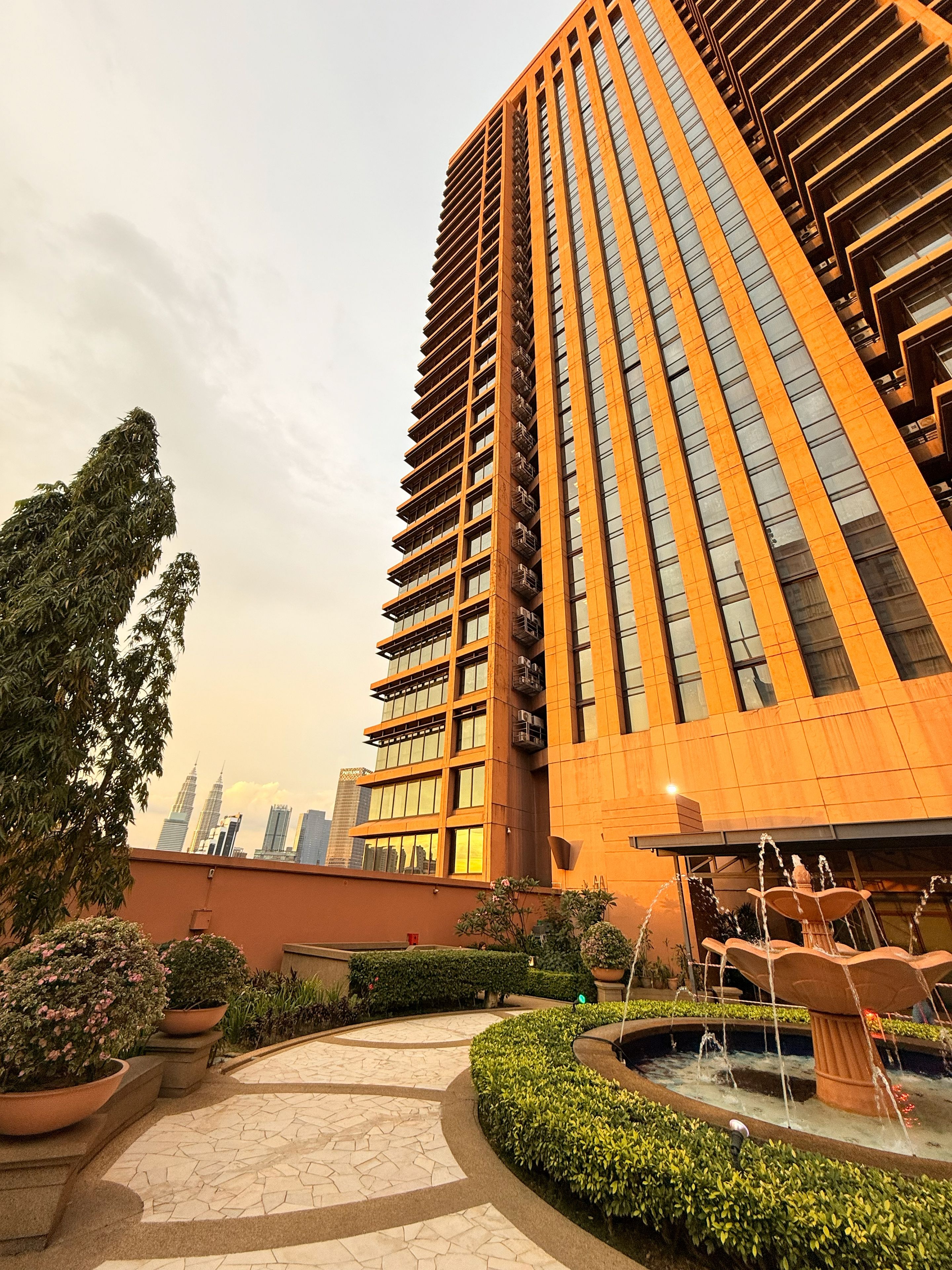 Un edificio moderno de gran altura con un diseño vertical llamativo, rodeado de jardines ajardinados y un elemento acuático.