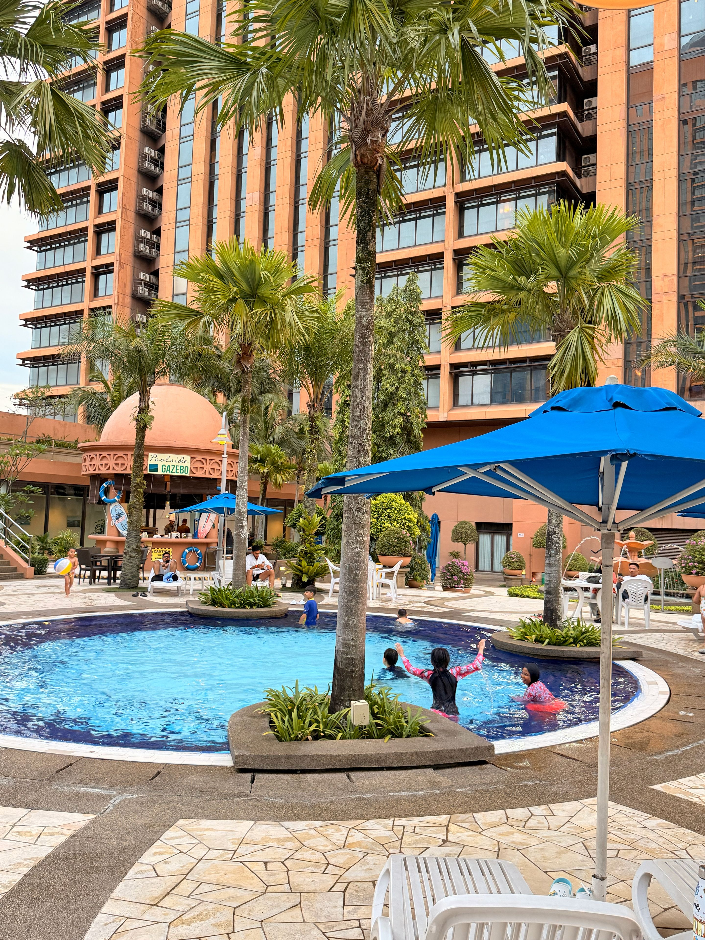 Zona de piscina de un hotel con palmeras, sillas de descanso y un edificio grande al fondo.