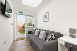 Een moderne kamer met een grijze bank, een bureau en een aan de muur bevestigde televisie. Boven de deur hangt een bord met daarop het vluchtroute-teken en aan de muur hangt een ingelijst kunstwerk.