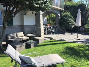Een terras met een bank, stoelen en een tafel, omringd door een verzorgd gazon en een stenen muur.