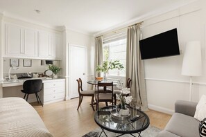 A modern living room with a sofa, a dining table with chairs, a TV mounted on the wall, and a kitchen area with cabinets and a window. - New! Cosy Chelsea Studio | Near Tube & King’s Road (Greater London)