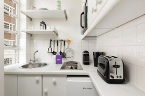 A modern kitchen with white cabinetry, a built-in stove, a toaster, and hanging utensils. - New! Cosy Chelsea Studio | Near Tube & King’s Road (Greater London)