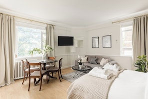 A hotel room with a bed, a sofa, a dining table with chairs, a TV, and a window with curtains. - New! Cosy Chelsea Studio | Near Tube & King’s Road (Greater London)