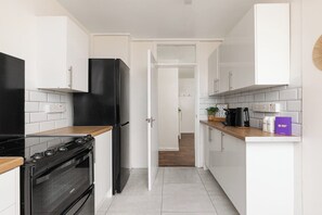 A narrow kitchen with white cabinets, a black refrigerator, a stove, and a microwave. - New! Light-Filled & Cosy 2BR Flat | Woolwich (Greater London)