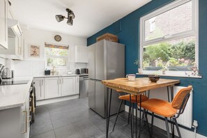 A modern kitchen with white cabinets, a stainless steel refrigerator, a wooden table with two chairs, and a window with a view of greenery. - New! Characterful 3BR Home | Garden & Parking (Greater London)