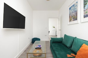 Ein Wohnzimmer mit einem grünen Sofa, einem Glastisch, einem an der Wand montierten Flachbildfernseher und gerahmten Kunstwerken.