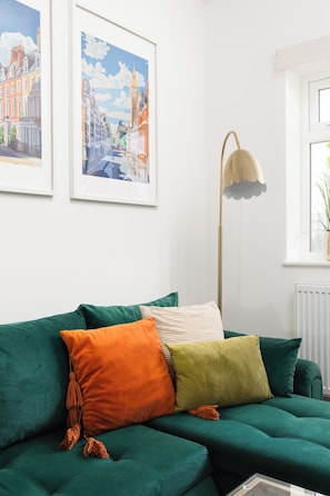 Ein Wohnzimmer mit einem grünen Sofa, orangefarbenen und grünen Kissen, einer Stehlampe und zwei gerahmten Stadtansichten an der Wand.