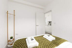 A hotel room with a bed, a green textured blanket, a wooden floor stand with a plant, and a white door leading to a bathroom. - New! Bright 2BR Flat near Plumstead Station (Greater London)