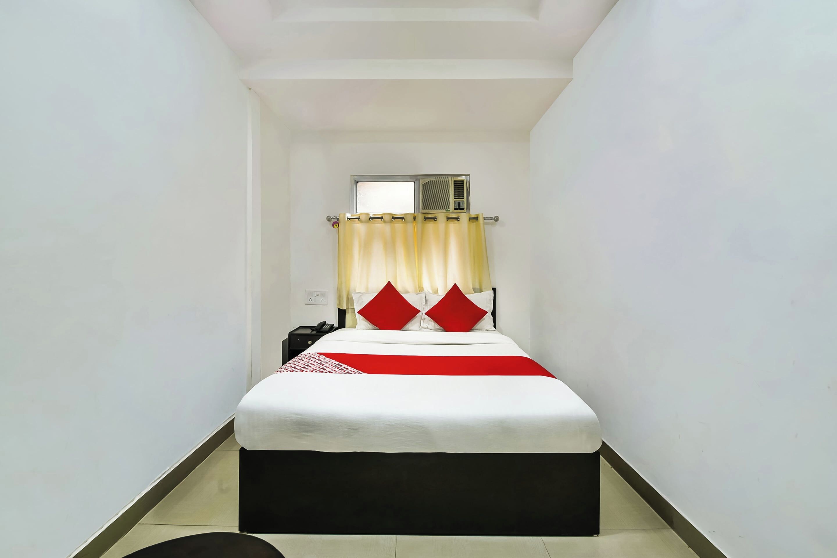 Habitación de hotel estrecha con cama, mesita de noche, teléfono y unidad de aire acondicionado en la pared.