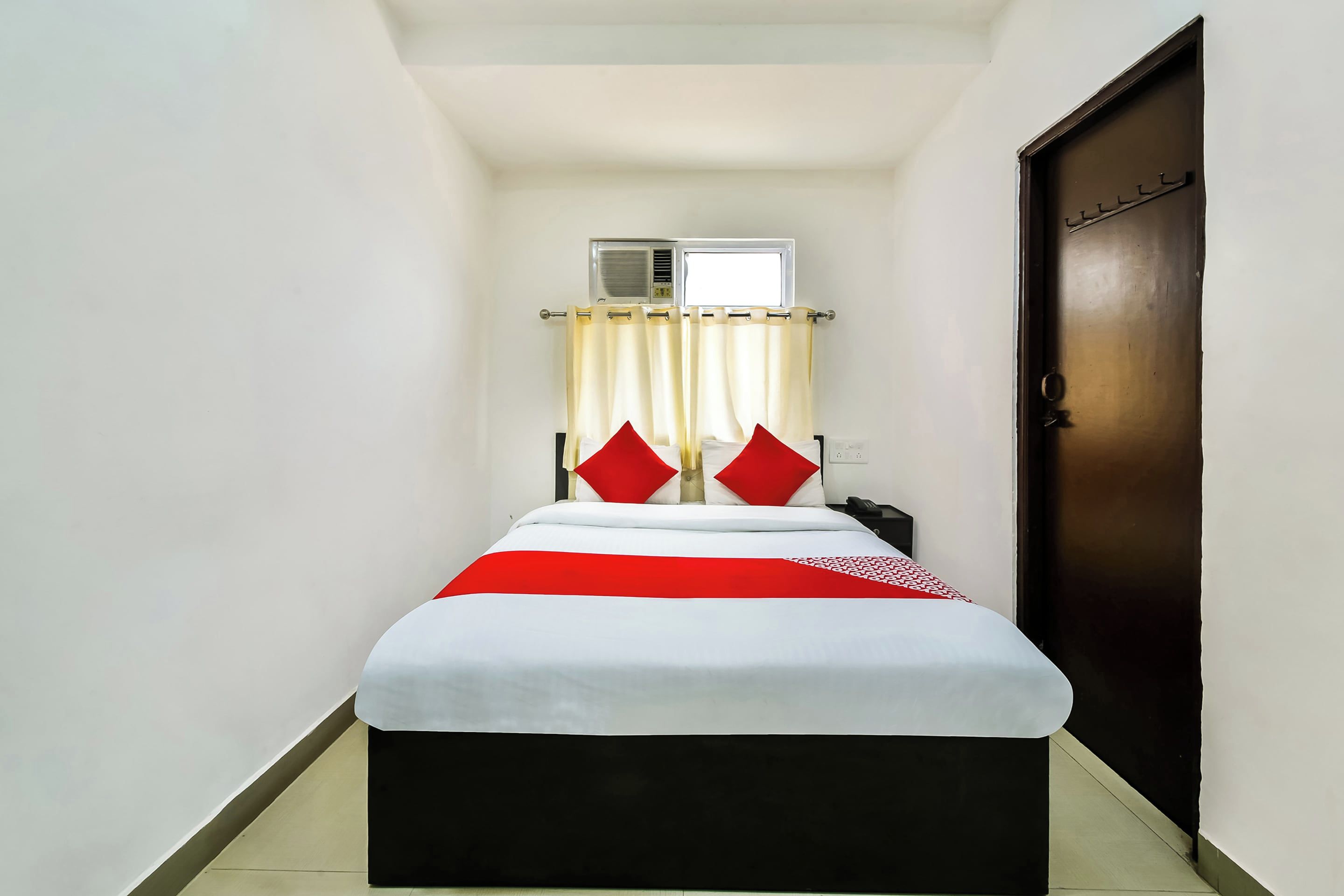 Una habitación de hotel pequeña y estrecha con una cama individual, una ventana con cortinas, una puerta y un equipo de aire acondicionado de pared.