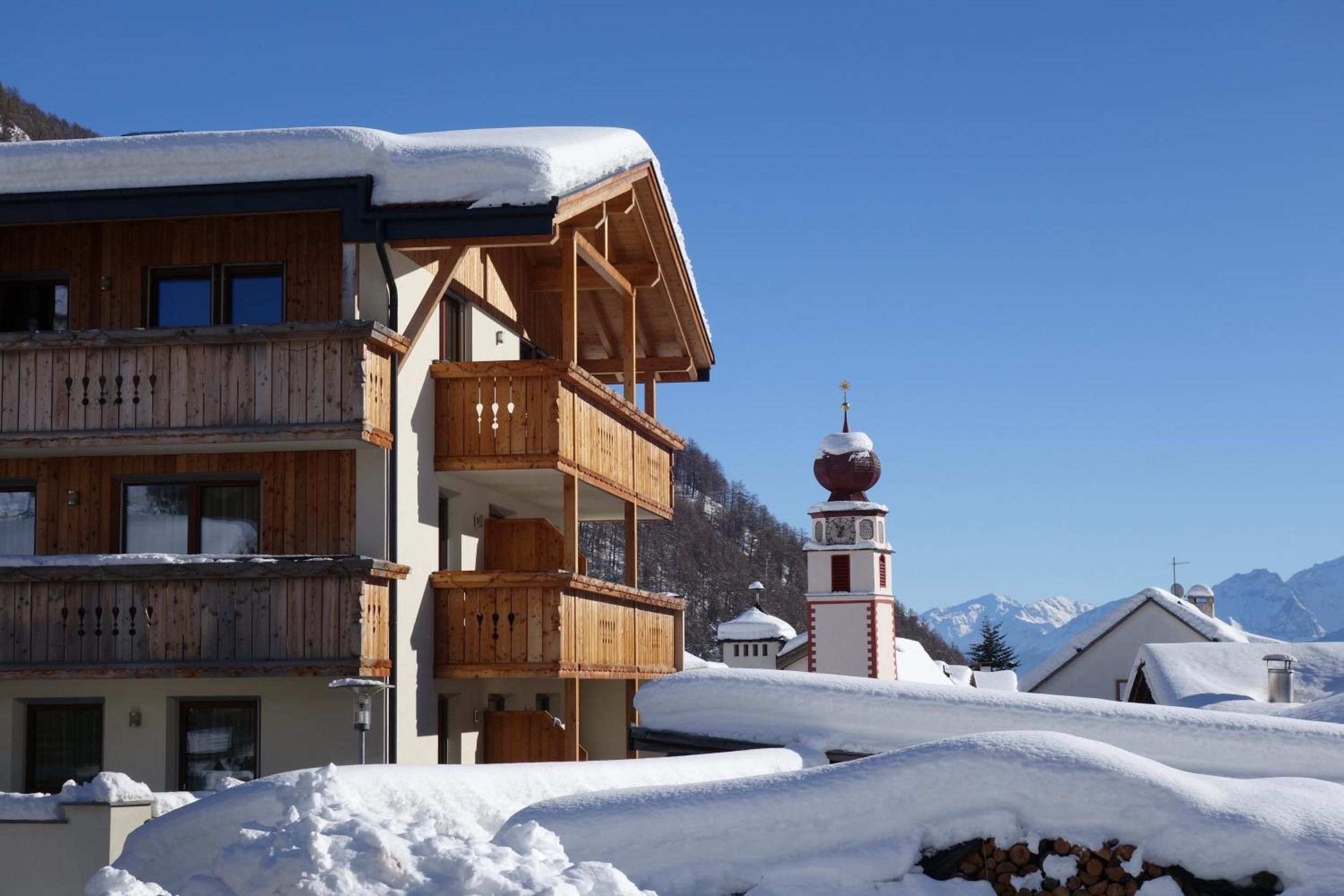 Una baita in legno con un balcone, una torre campanaria e montagne sullo sfondo.