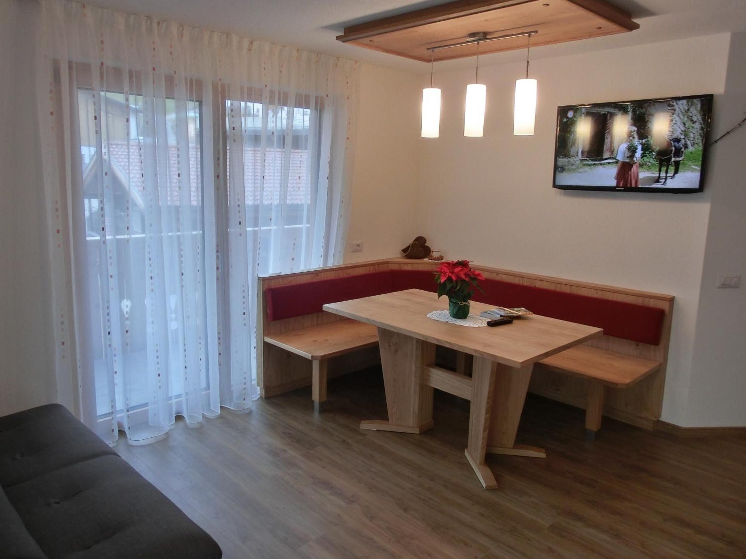 Ein Zimmer mit einer Sitzecke mit Holzbank, einem Tisch mit Blumenvase und einem an der Wand befestigten Fernseher.