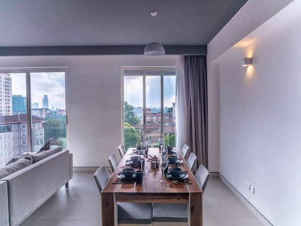 Un comedor moderno con una larga mesa de madera dispuesta para seis personas, un sofá y un amplio ventanal con vistas a la ciudad.