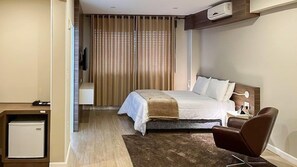 A hotel room with a bed, bedside table, chair, and a small refrigerator. - Imperatriz Hotel (Lajeado)