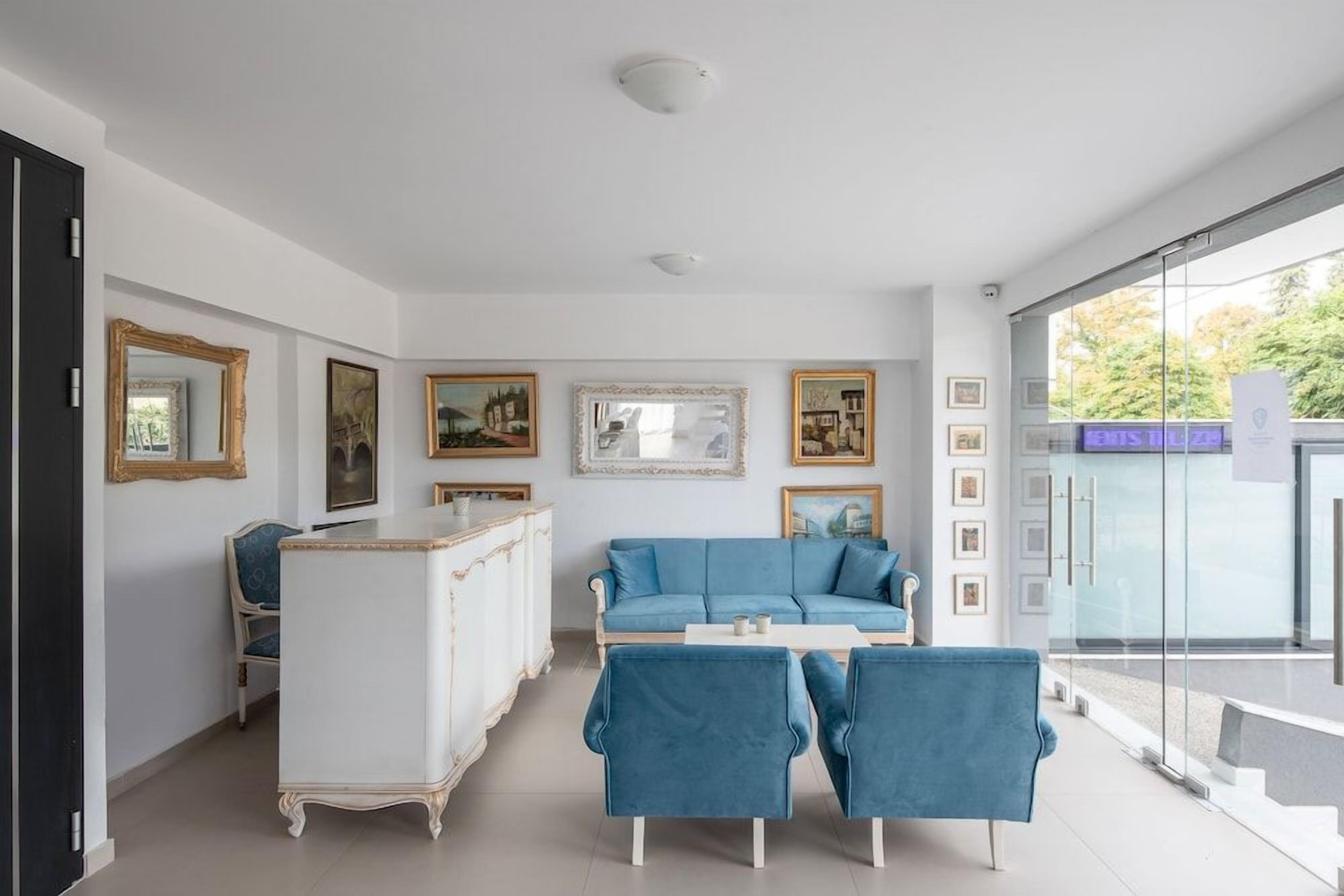 A modern living room with a blue sofa, two blue armchairs, a white coffee table, and a white cabinet. There are framed pictures on the walls and a glass door leading outside.