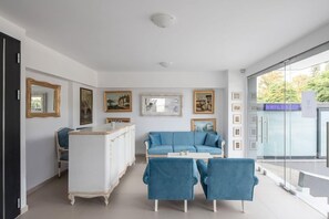 Uma sala de estar moderna com um sofá azul, duas poltronas azuis, uma mesa de centro branca e um armário branco. Há quadros nas paredes e uma porta de vidro que dá para o exterior.