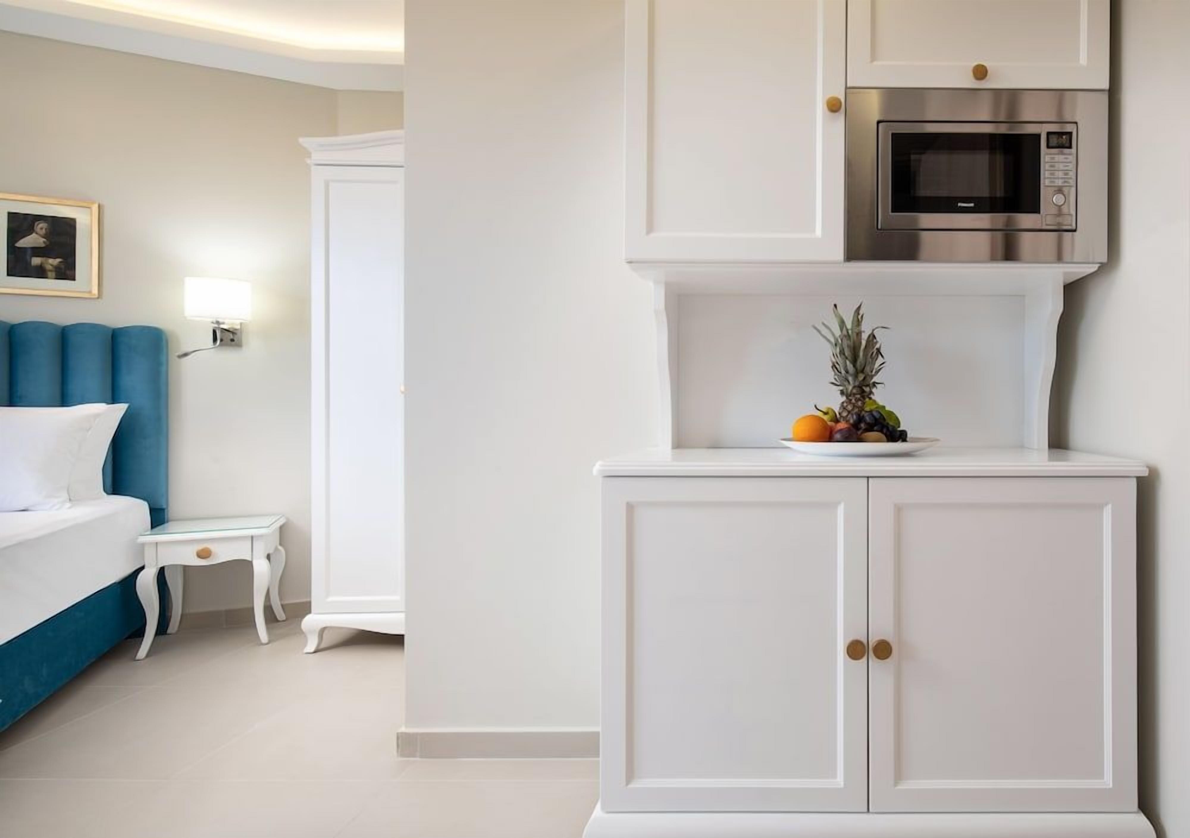 Habitación de hotel con cama, una mesita de noche, un armario blanco con microondas y una estantería en la pared con frutas.