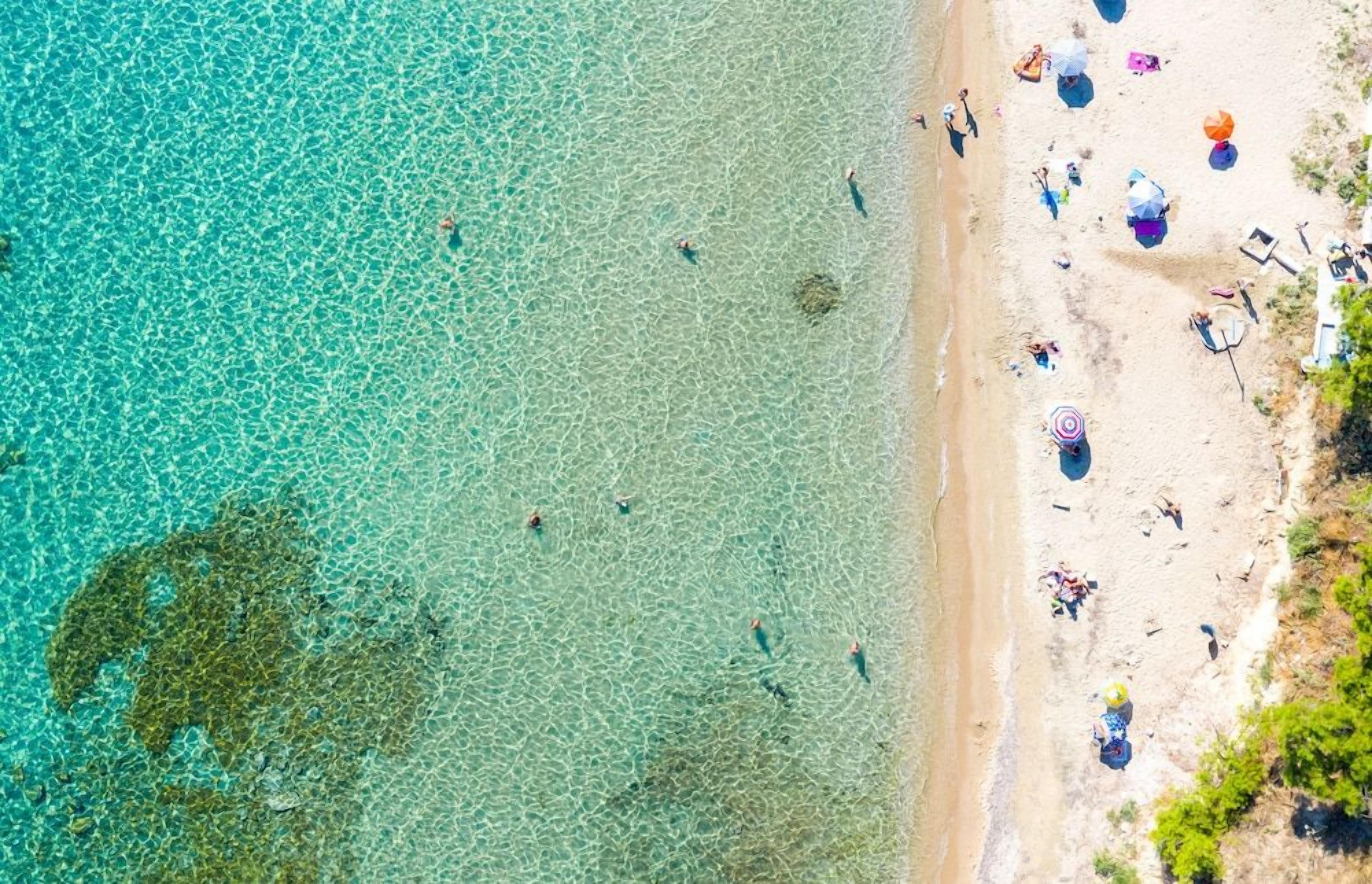 Une vue aérienne d’une plage aux eaux turquoise et limpides, où l’on voit des gens se baigner et des parasols plantés dans le sable.