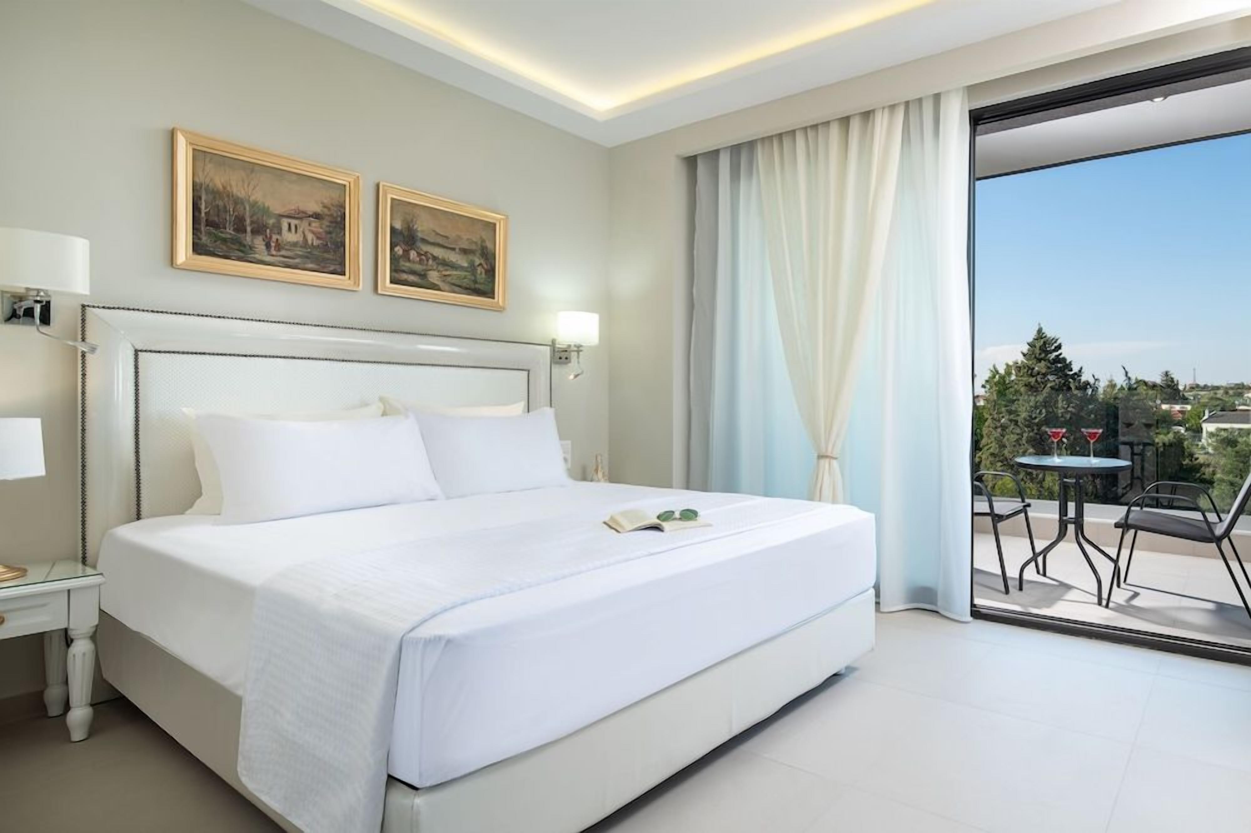 Habitación de hotel con una cama grande, una mesita de noche, una lámpara, dos cuadros en la pared y vistas al exterior a través de un ventanal.