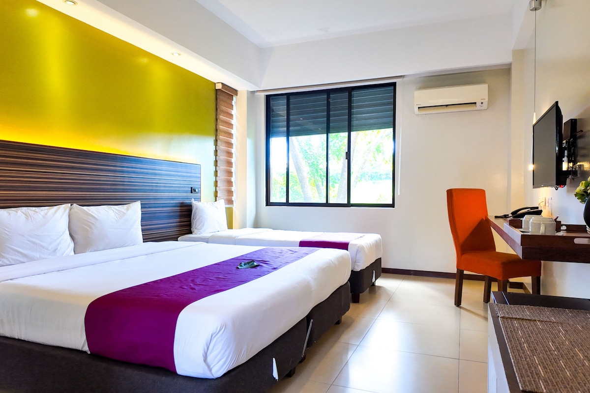 A hotel room with two beds, a desk with a chair, a TV, and a window with a view of greenery.