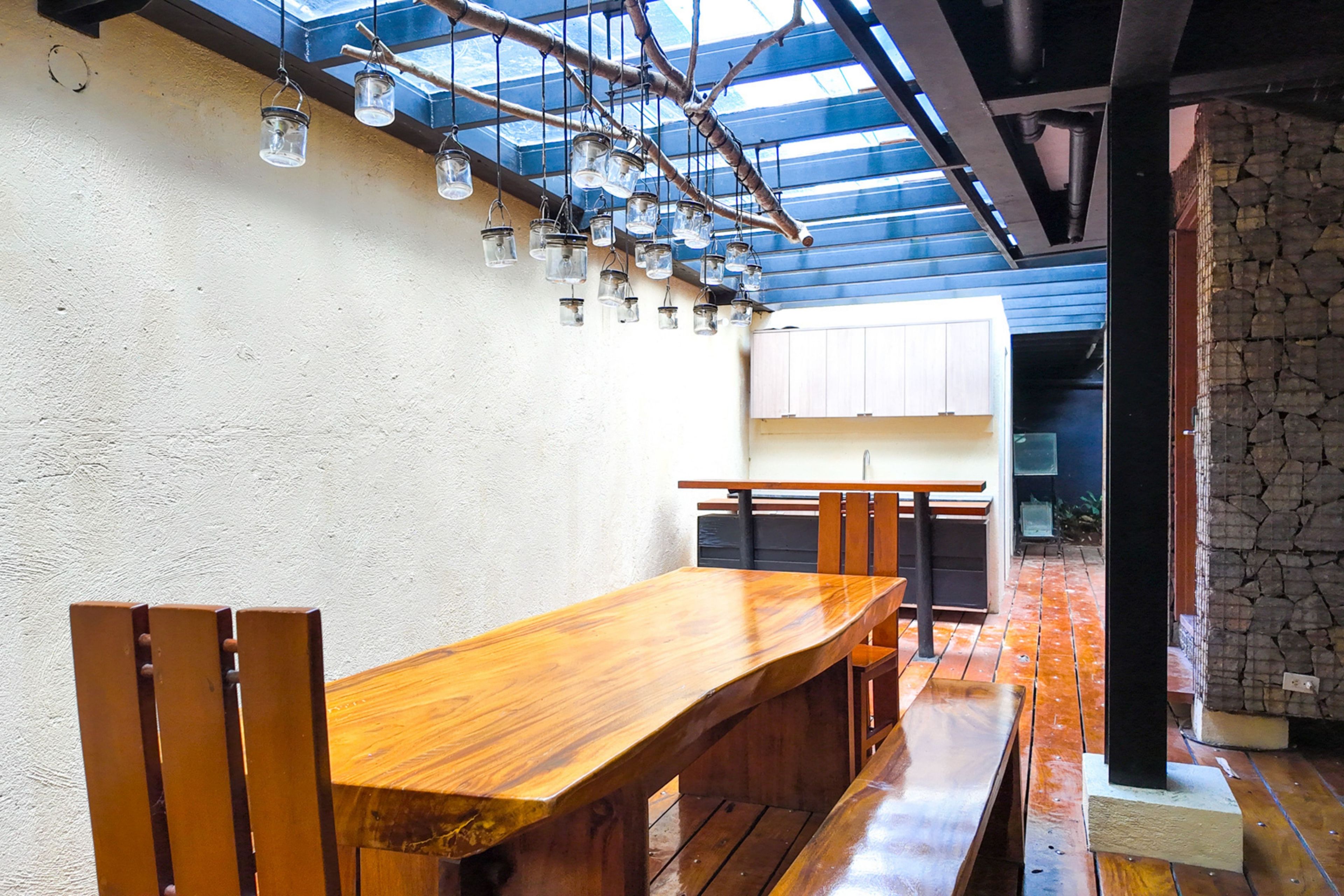 Une table à manger en bois avec des bancs, un coin cuisine doté de meubles blancs et un mur en pierre.