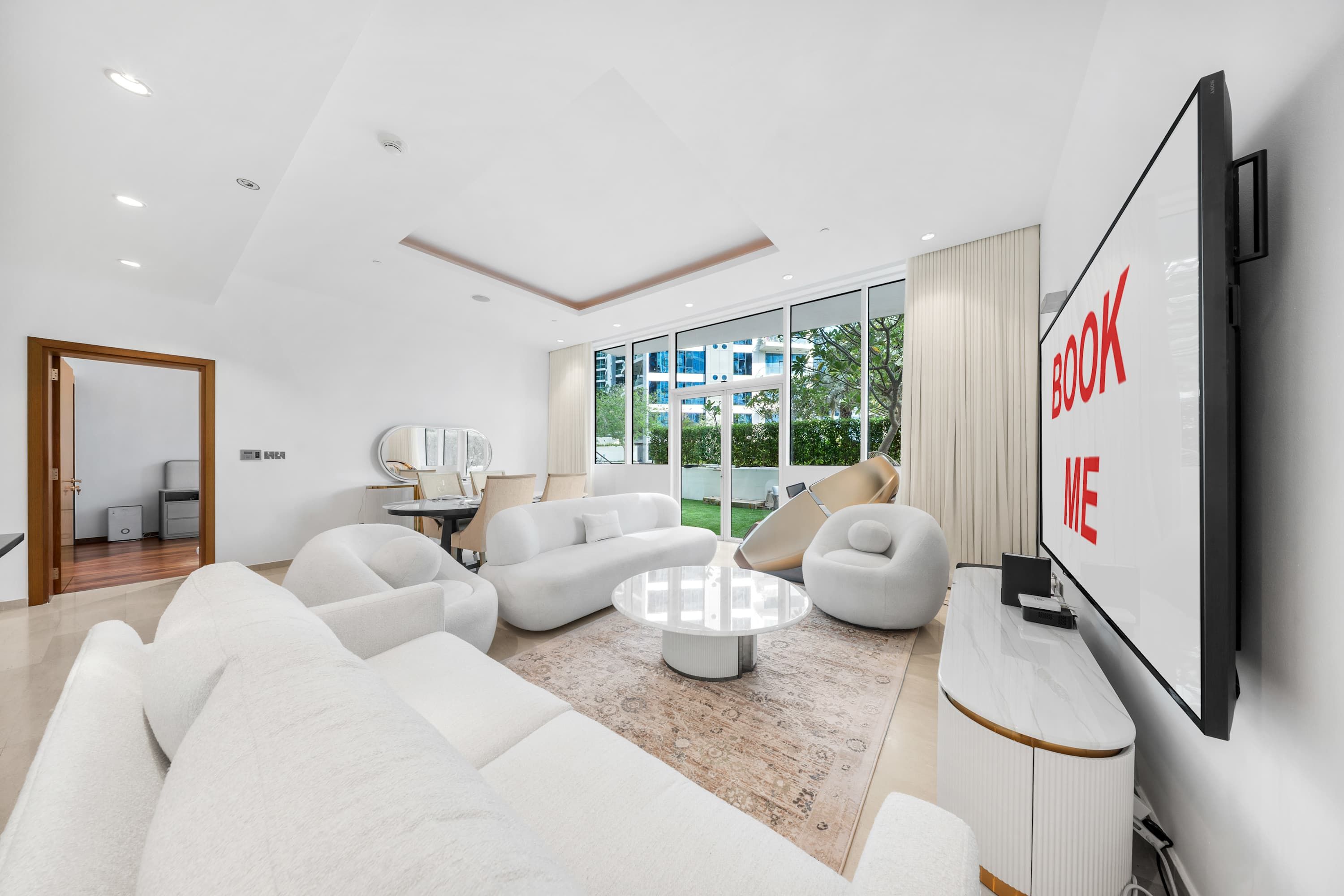 A modern living room with a large flat-screen TV, white sofas, a coffee table, and a view of greenery outside.