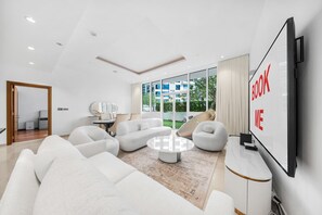 Ein modernes Wohnzimmer mit großem Flachbildfernseher, weißen Sofas, einem Couchtisch und Blick auf Grünflächen.