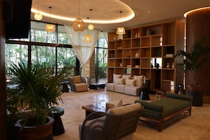 Amplia sala de estar con sofá, sillones y una mesa de centro. Cuenta con ventanas amplias con cortinas, una estantería y plantas en macetas.