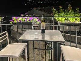 Una mesa con una botella de vino y una copa, rodeada de sillas blancas, con plantas en macetas y una barandilla al fondo.