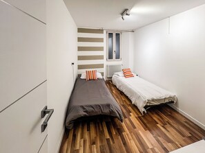 Habitació petita amb dos llits individuals, un radiador, una finestra amb cortines i un terra de fusta.
