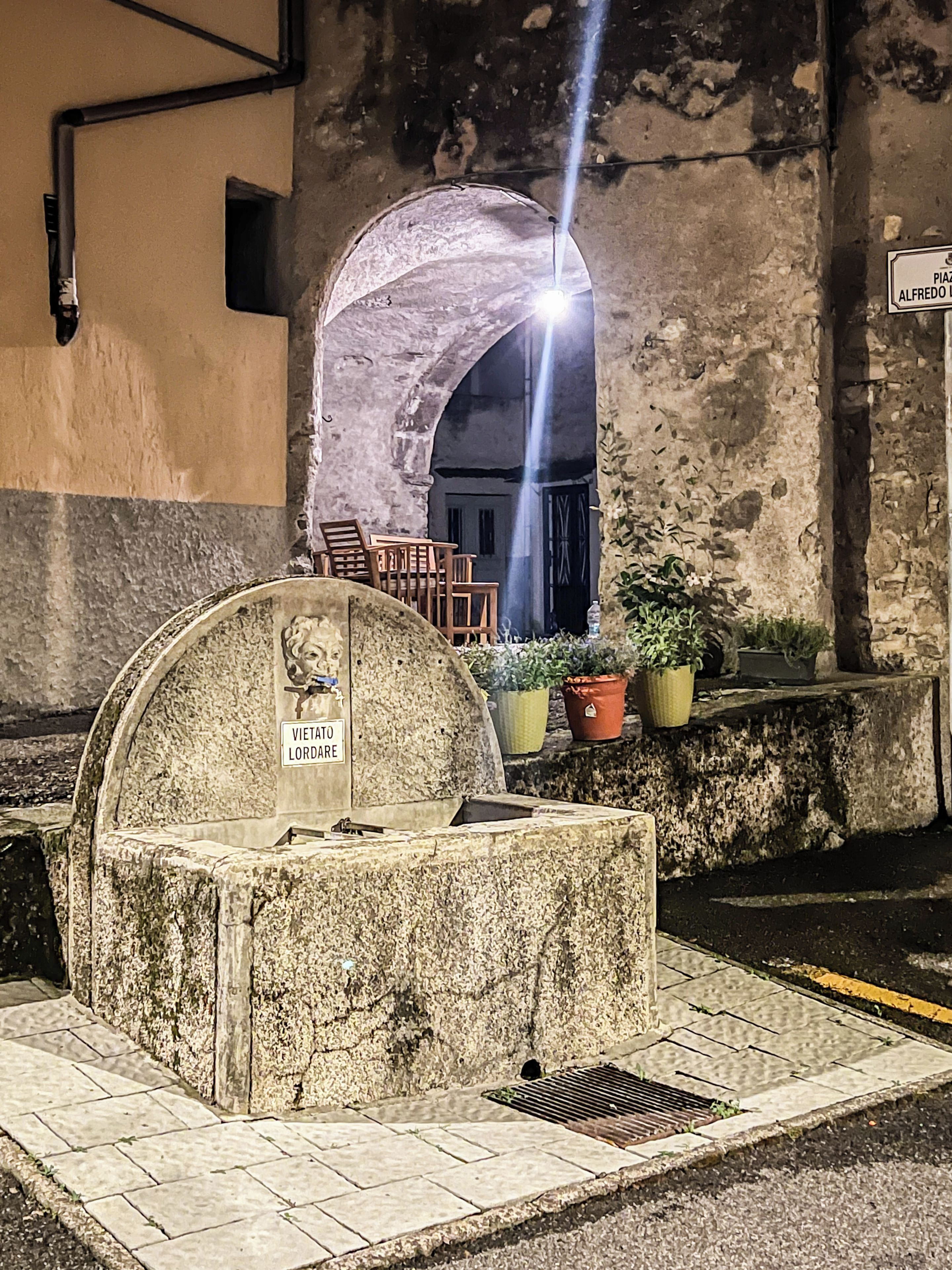 Un monumento de piedra con una placa, un banco y plantas en macetas en un entorno urbano.