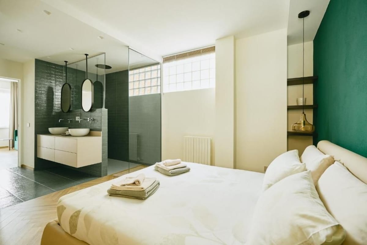 A modern bedroom with a large bed, a glass-enclosed bathroom, and a view of a cityscape.