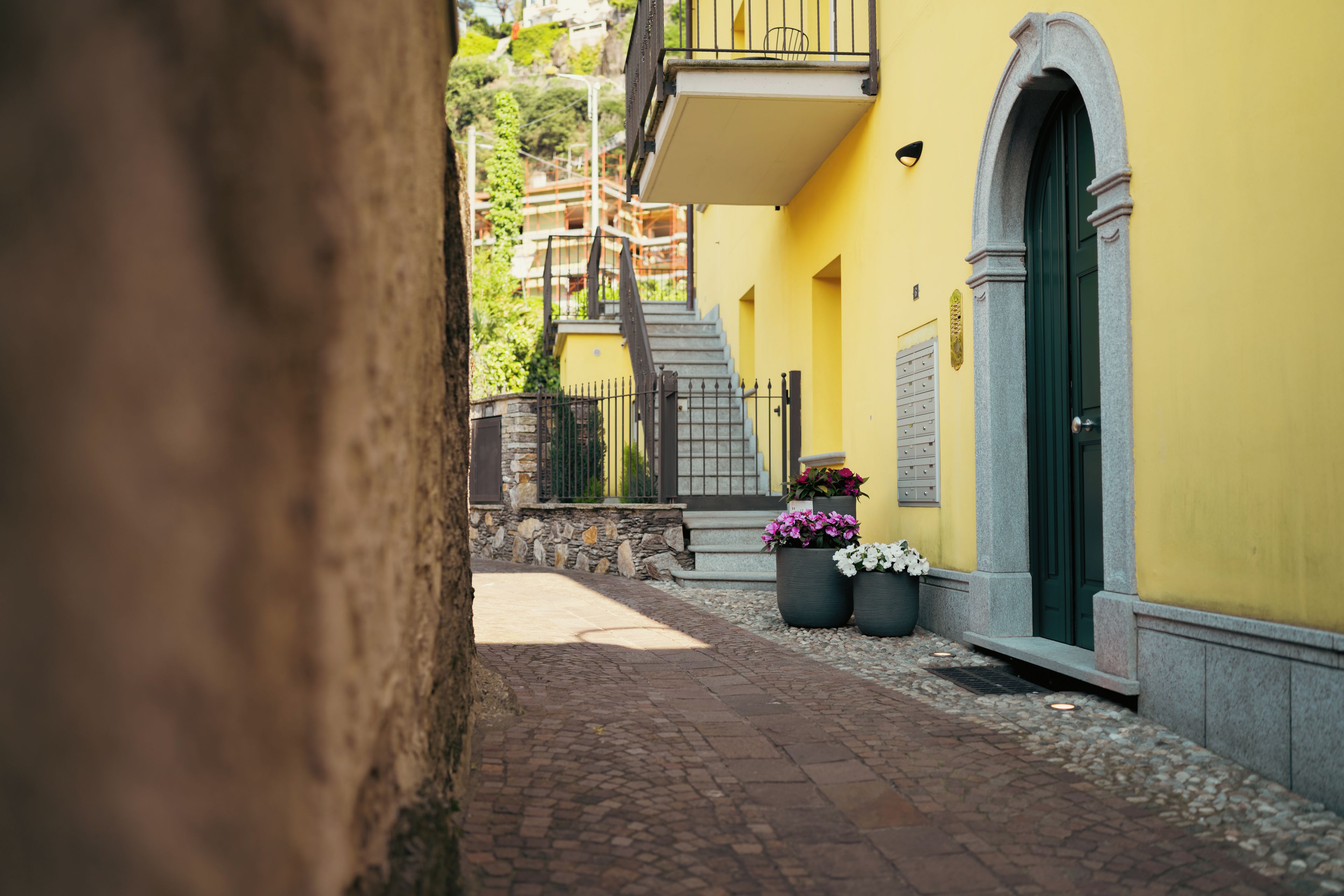 A narrow alleyway with a yellow building, a stone wall, and a green door. There are potted plants and a staircase leading to an upper level.
