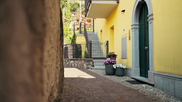 A narrow alleyway with a yellow building, a stone wall, and a green door. There are potted plants and a staircase leading to an upper level.