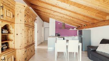 A modern kitchen with white cabinets and a purple accent wall, a dining area with white chairs, and a dark sofa.