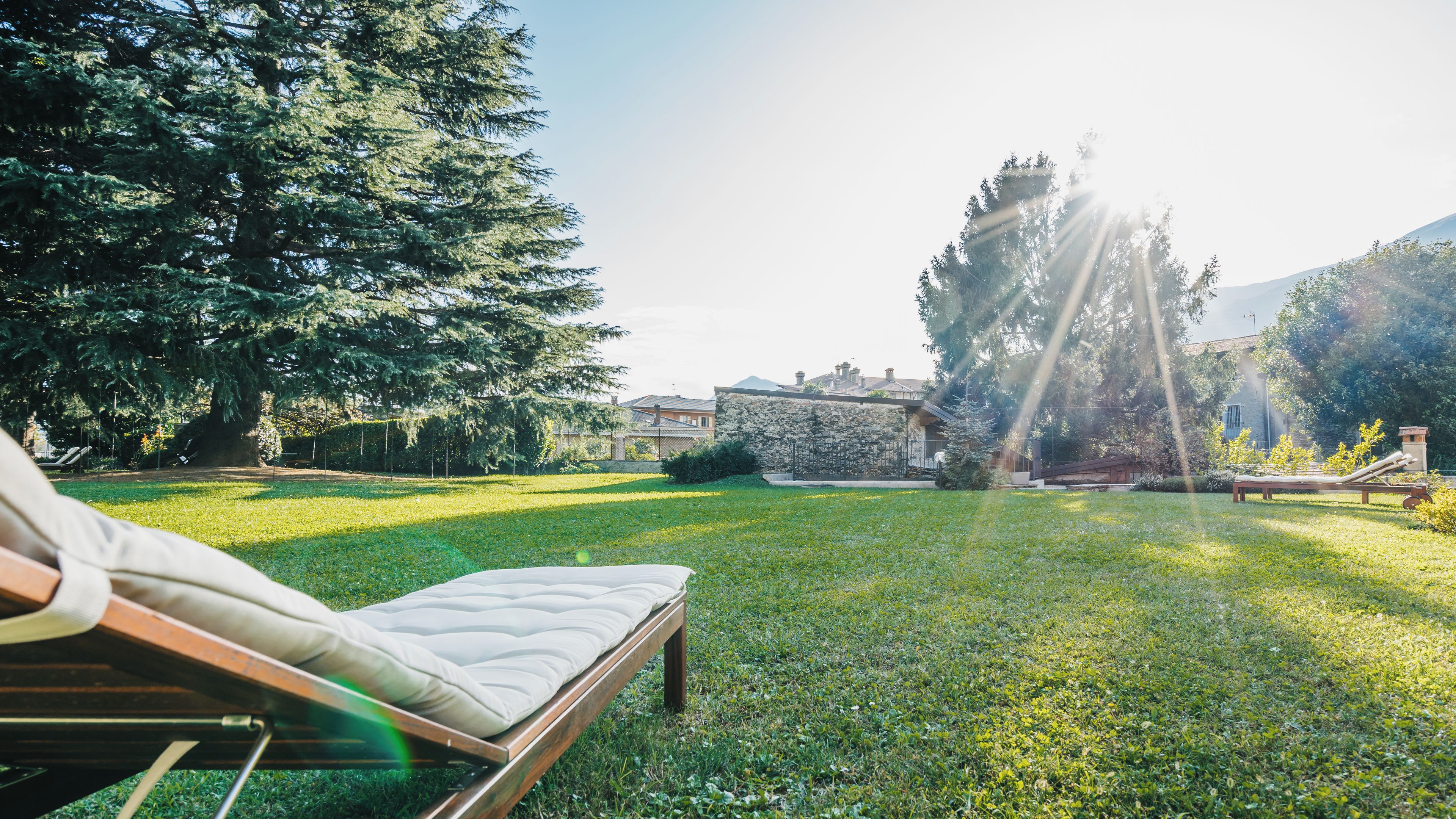 A grassy outdoor area with a lounge chair and a mattress, surrounded by trees and a stone wall.