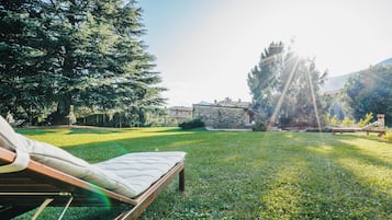A grassy outdoor area with a lounge chair and a mattress, surrounded by trees and a stone wall.