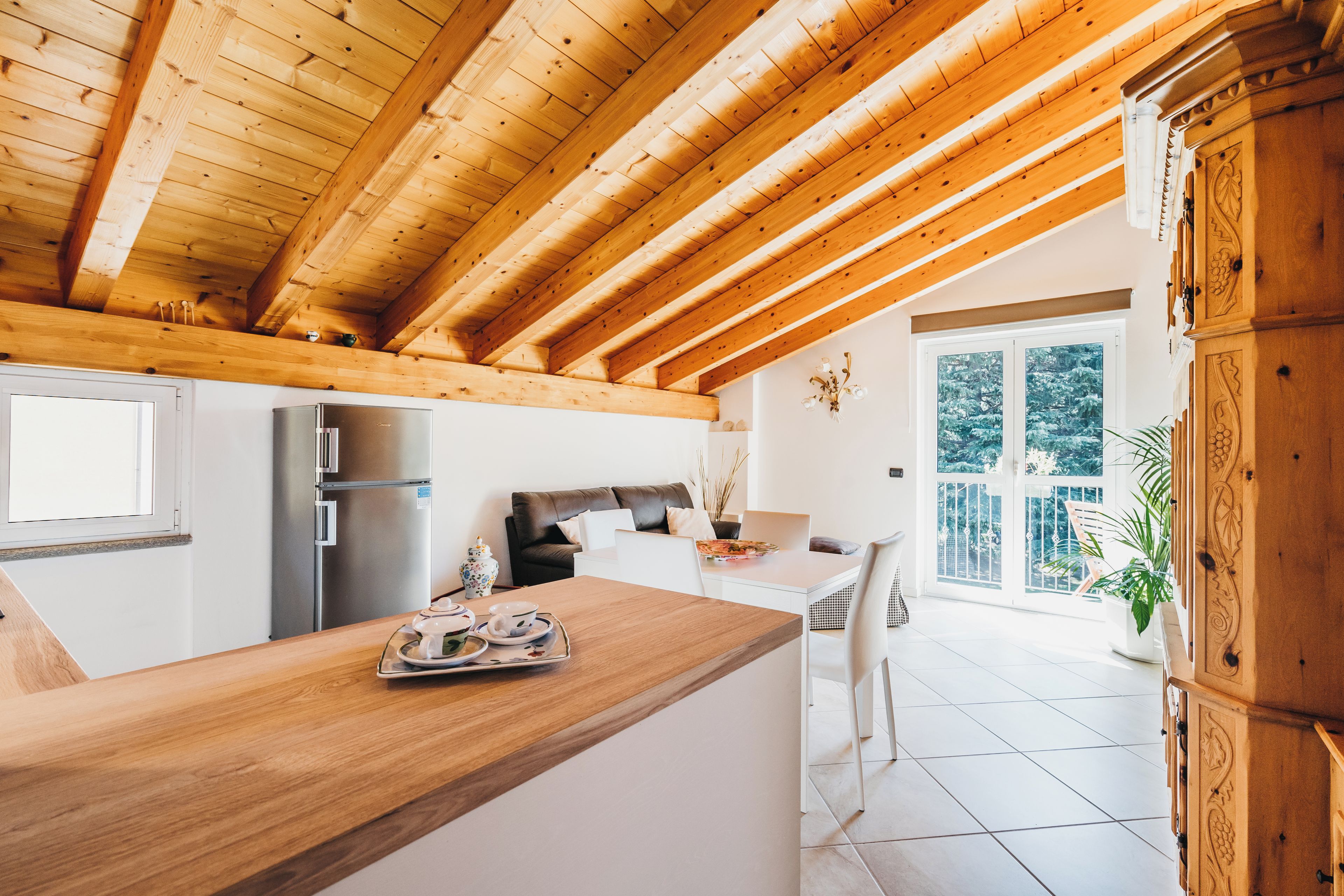 A modern kitchen with wooden beams, a stainless steel refrigerator, and a dining area with a view of the outdoors.
