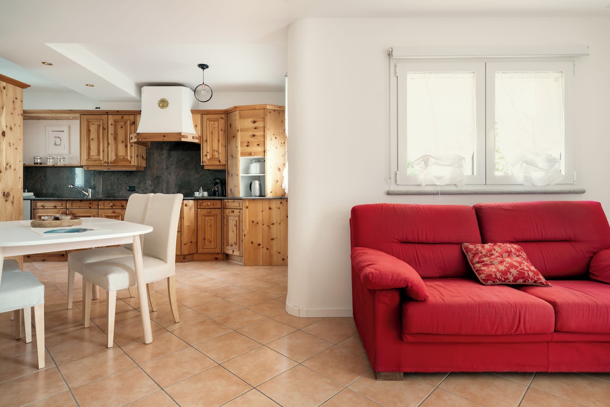 A modern kitchen with wooden cabinets, a dining area with a white table and chairs, and a red sofa with a decorative pillow.