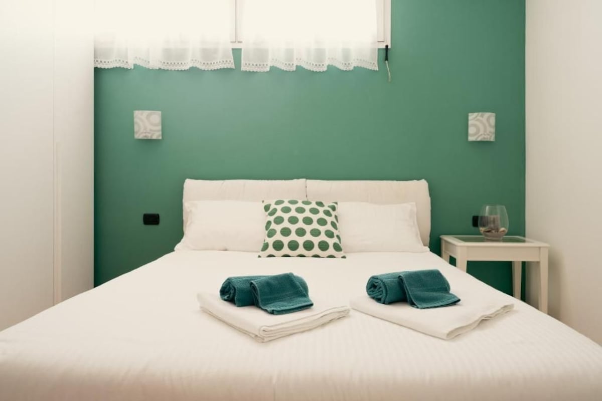 A neatly made bed with two folded towels, a green polka dot pillow, and a small bedside table with a glass and a decorative item.