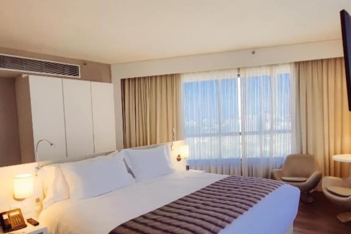 Una habitación de hotel con una cama grande, mesitas de noche, una silla y un ventanal con cortinas.
