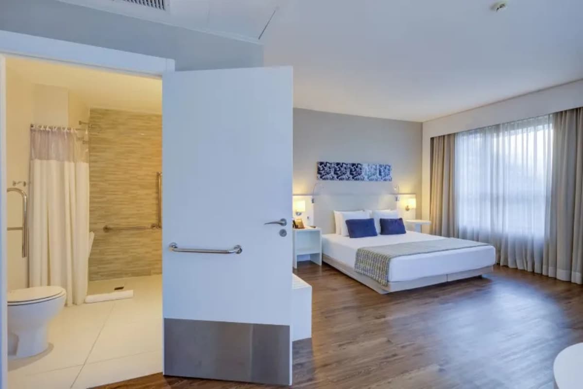 Habitación de hotel con ducha, cama, mesita de noche y un gran ventanal con cortinas.
