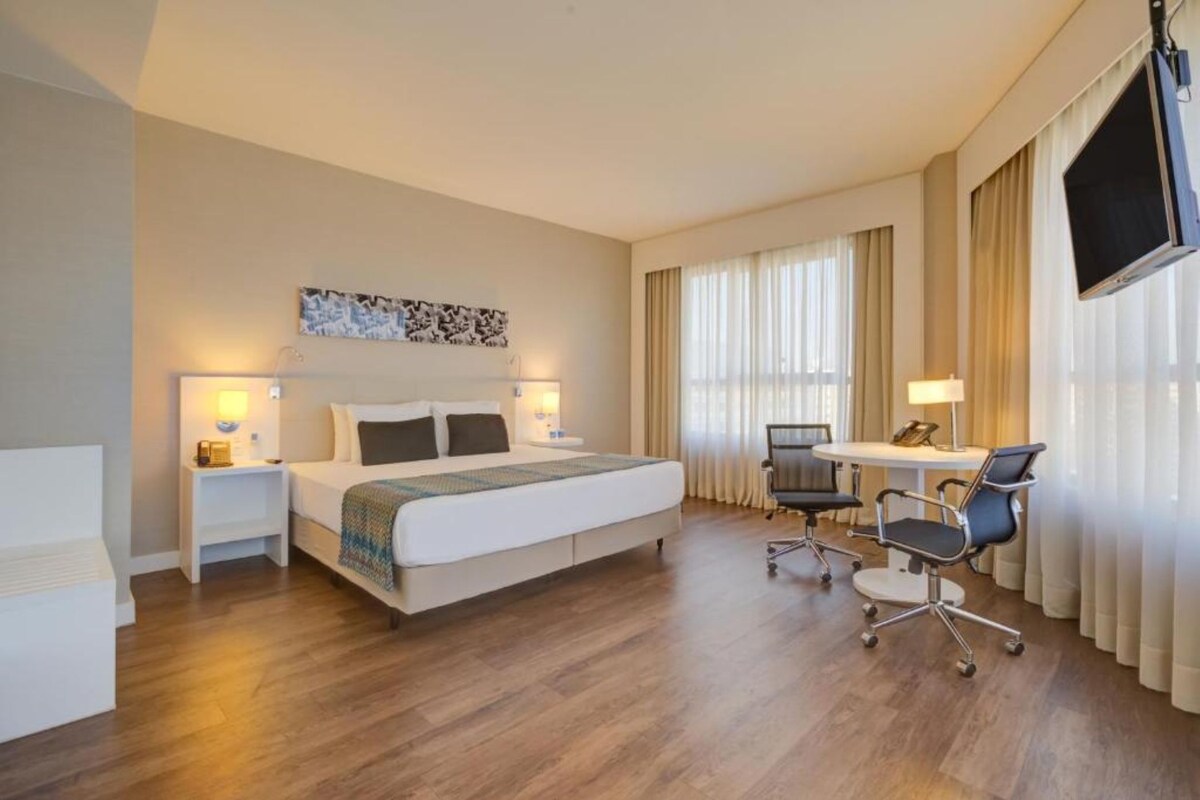 Una habitación de hotel con cama, mesitas de noche, escritorio con silla y televisor montado en la pared.