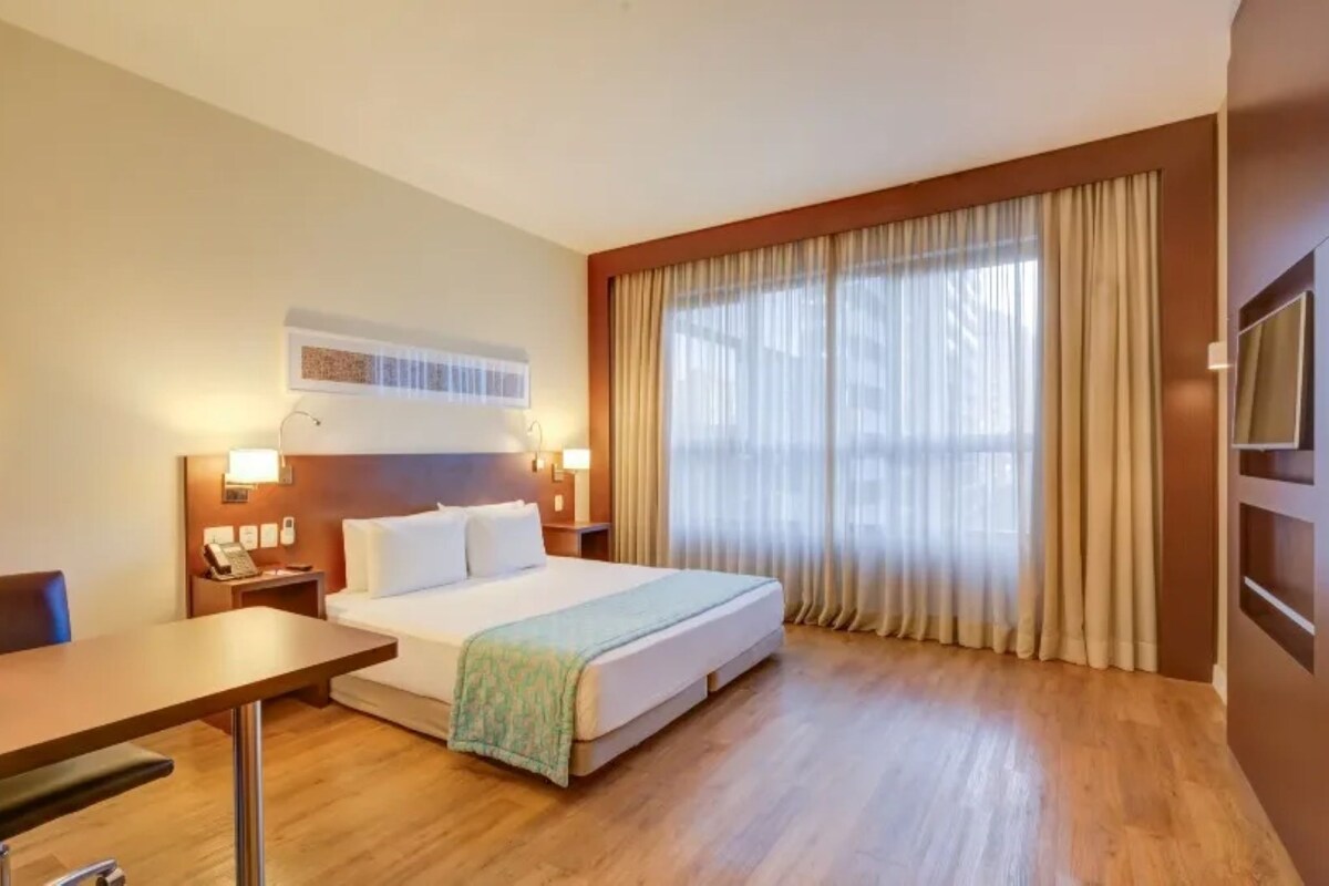 Una habitación de hotel con cama, mesitas de noche, un escritorio y un ventanal con cortinas.