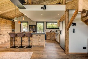 A modern kitchen with wooden accents, a bar area with stools, and large windows. - Beautiful chalet in heart of Chamonix sleeps 8-10, 4 bed/4 bath (+king sofa bed) (Chamonix-Mont-Blanc)