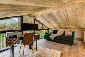 A cozy room with a wooden ceiling, a desk with a computer, a green sofa, and a large window offering a view of houses and trees. - Beautiful chalet in heart of Chamonix sleeps 8-10, 4 bed/4 bath (+king sofa bed) (Chamonix-Mont-Blanc)