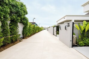 A clean, modern pathway flanked by neatly trimmed hedges and a row of compact, white buildings with visible door numbers. - New Modern 2BR Pool Villa in Peaceful Rawai (Tambon Rawai)
